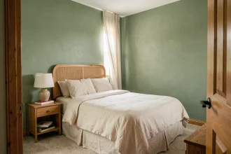 Petite chambre adulte peinte en vert sauge mat avec literie en lin ivoire et tête de lit en rotin, illustrant comment une teinte douce agrandit visuellement l'espace
